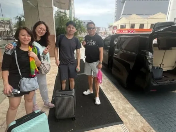 A group with luggage beside a black MPV on a city street with heritage shophouses