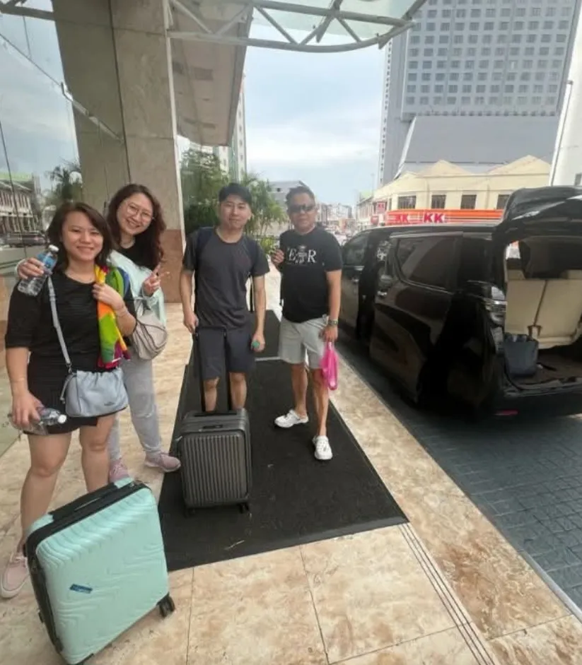 A group with luggage beside a black MPV on a city street with heritage shophouses