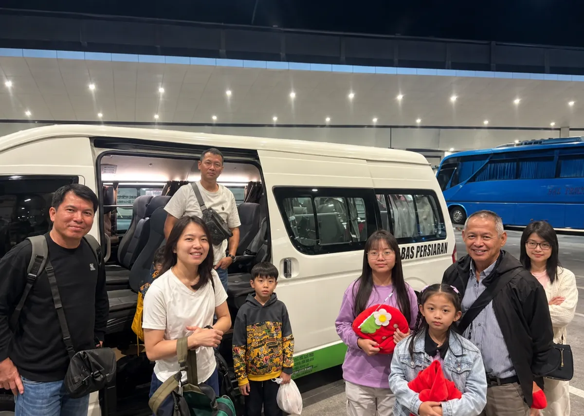 A family holding flower toys beside a Bas Persiaran tour bus at night