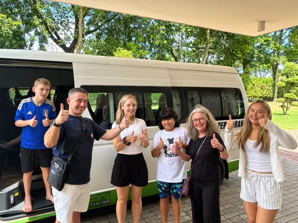 An international family giving thumbs up beside a Persiaran tour bus in a park setting